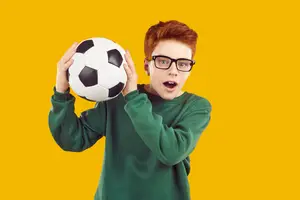A young boy with glasses holds a soccer ball with both hands and seems to be posing for a photo with a surprised expression