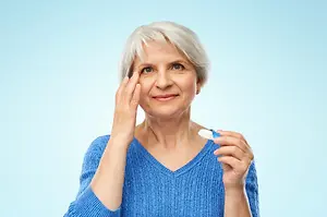 An older woman with white hair is holding a toothbrush and applying something to her eye while smiling.