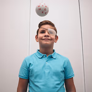 A boy wearing a turquoise shirt and eye patch smiles while looking at a ball with letters, against a plain background.