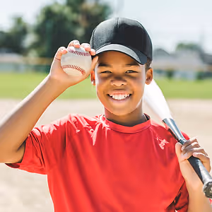 Smiling boy in a red shirt and black cap holding a baseball and bat on a sunny day.