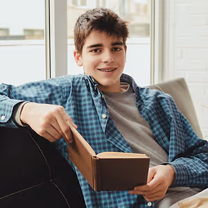 Smiling young person with braces reading a book on a sofa by a window.