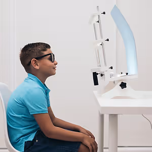 A boy in a blue shirt and glasses sits at a table with a vision testing device.