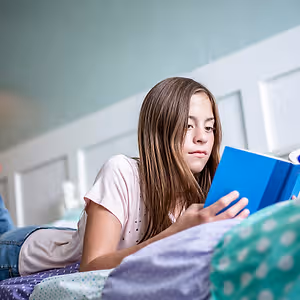 A girl lying on a bed reading a blue book in a softly lit room.