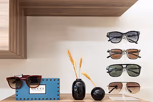 A wooden shelf displays several pairs of sunglasses, including Gucci and Tom Ford brands, along with decorative items such as a blue sign, a wheat stalk in a vase, and a red pair of sunglasses on top of a blue box.