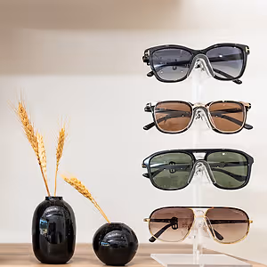 Four pairs of sunglasses are displayed on a stand next to a vase with wheat, showcasing a collection of shades in various colors and styles, against a neutral background.