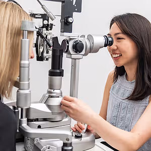 A woman with long dark hair is smiling and having her eyes examined with a slit lamp in a clinic setting.