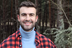 A man with a beard is smiling and posing for a photo in a forest area