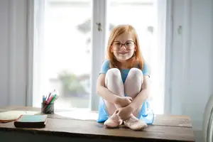 A young girl with glasses and ballet shoes is sitting on a table in a room with a glass window and sunlight shining on her