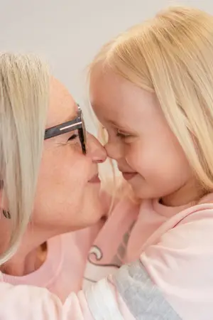 An adult woman wearing glasses is embracing a young girl with blond hair, both are dressed in pink clothing, the woman is wearing a gray earring, and they are standing in a room with white walls.