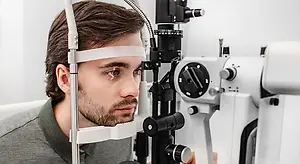 A man wearing glasses sits in front of an eye exam machine and looks into it