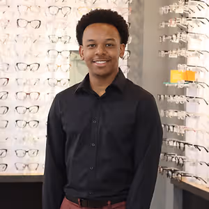 A smiling man in a black shirt stands in front of a wall of glasses