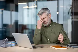 An older man in a green shirt sitting at a desk rubbing his forehead with one hand and holding his glasses with the other.