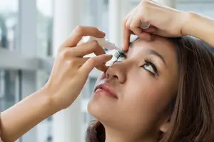 A woman applies eye drops while looking in a mirror.