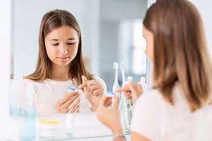 Two women standing in front of a mirror, one brushing her teeth with a toothbrush.