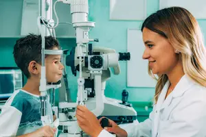 A woman in a white lab coat examining a young boy's eyes with an ophthalmoscope.