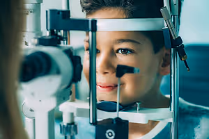 A child wearing a blue shirt is smiling while looking through an ophthalmoscope in an eye clinic.