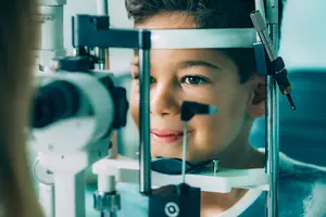 A child wearing a blue shirt is smiling while looking through an ophthalmoscope in an eye clinic.