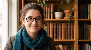 A woman with glasses and a scarf sits in front of a bookshelf with books and plants