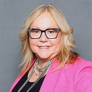 Portrait of a woman with blonde hair, wearing glasses, a pink blazer, and a necklace