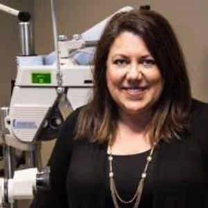 A woman wearing a black top and necklace is smiling for a picture while standing in a room with a medical machine behind her.