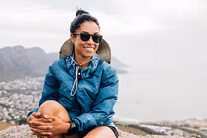 A woman wearing a blue jacket, sunglasses, and a hat is sitting on a rock and smiling while looking at the camera. Behind her, there is a view of a mountain and a city.