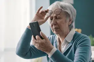 An elderly woman adjusts her glasses while looking at her phone.