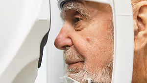 An older man wearing glasses is looking through a white eye exam device in a room