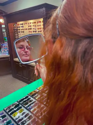 A woman wearing glasses is looking at herself in a mirror in a shop.