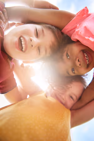 Three young children, one boy and two girls, are smiling and appear to be posing for a photograph with their heads together in a circle, and the sunlight is shining on them.