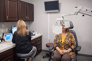 A woman sits in an optometrist's chair getting her eyes checked while another woman sits at a computer in the background.