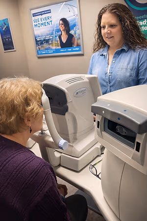 An adult woman with blonde hair is using a Topcon machine to check her eye sight while another woman is standing behind her.