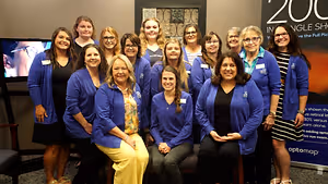 Twelve women wearing blue jackets with the text '200' in the background standing in front of a television and a banner