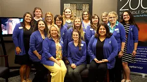 Twelve women wearing blue jackets with the text '200' in the background standing in front of a television and a banner