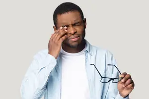 A man with his eyes closed and his hand on his forehead, holding a pair of glasses in his other hand.