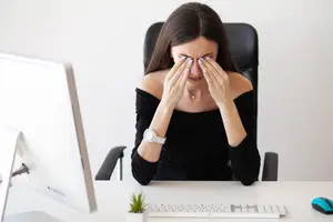 A woman sitting at a desk with her hands on her face, possibly experiencing discomfort or pain. She is wearing a black long-sleeve shirt and a watch. She is sitting in front of a computer monitor and a keyboard, with a potted plant on the desk. She is in an office setting, possibly working or feeling stressed.