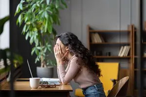 A woman sitting at a desk with her hands covering her face, looking worried or anxious.