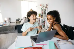 A boy and a woman are sitting in front of a desk with a laptop and books, smiling and laughing.