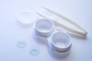 An empty contact lens case with two lenses in it, along with a pair of tweezers and two transparent containers on a white surface.