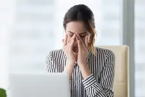 A woman sitting in an office chair is covering her eyes with her hands, seemingly in pain.