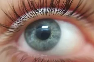 Close-up of a person's eye with long lashes and a slightly open mouth, the iris appears to be light gray with a darker gray pupil, and the eyelashes are thick and curled.