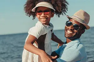 A man wearing a blue shirt and a hat is smiling while carrying a child on his shoulders on a beach. The child is wearing a white dress and sunglasses. Behind them, the sea is calm and the sky is clear.