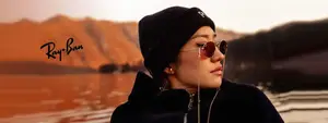 East Asian woman in a black coat, hat, and Ray-Ban sunglasses poses for a photo in a desert landscape with mountains and water in the background.