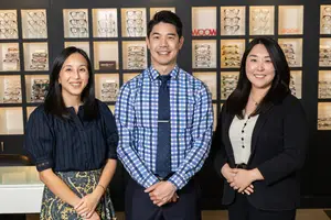 Three smiling adults, two women and one man, standing in front of a display of glasses