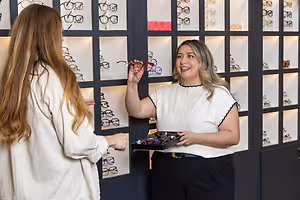 Two women smiling at each other, one holding a red-framed pair of glasses, inside a shop with shelves of glasses in the background.