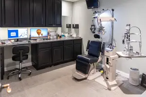 An eye examination room with a chair, desk, computer monitors, cabinets, and medical equipment.