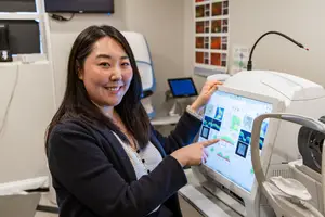 A woman is standing and pointing at the monitor of an eye exam machine