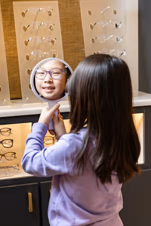 A girl is holding a mirror and looking at herself in a store that has shelves with glasses frames displayed on them