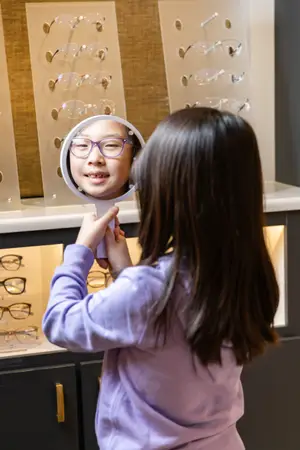 A girl is holding a mirror and looking at herself in a store that has shelves with glasses frames displayed on them