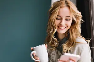 A woman smiles and holds a white mug while looking at her phone