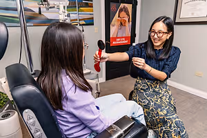 A woman and a young girl are in an optical clinic, with the woman demonstrating something to the girl while the girl looks at her with a curious expression. The room has a black door with a poster on it, and a white wall with a certificate and a painting.